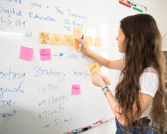 A woman with long hair is writing on a whiteboard. She is using sticky notes and markers to organize and plan a strategy. The board has various handwritten notes and symbols related to marketing and digital education.