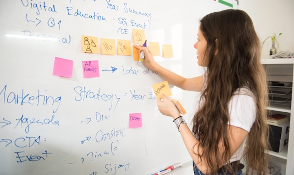 A woman with long hair is writing on a whiteboard. She is using sticky notes and markers to organize and plan a strategy. The board has various handwritten notes and symbols related to marketing and digital education.